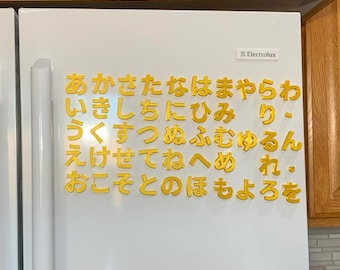 May include: A white refrigerator door featuring gold-colored Japanese hiragana characters arranged in a grid. The Electrolux brand name is printed in the upper right corner. The characters are organized in rows and columns, creating a decorative display.