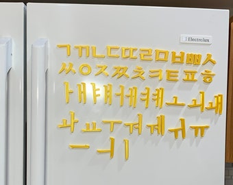 May include: A collection of yellow, three-dimensional Korean Hangul characters arranged on a white refrigerator door. The set includes various consonants and vowels, each featuring a raised, block-like structure that adheres to the smooth surface.