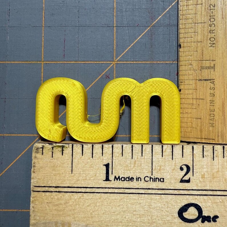 May include: A bright yellow, three-dimensional plastic letter 'm' positioned against a wooden ruler. The letter has a textured surface and measures approximately 4.5 centimetres in width, as indicated by the markings on the ruler.