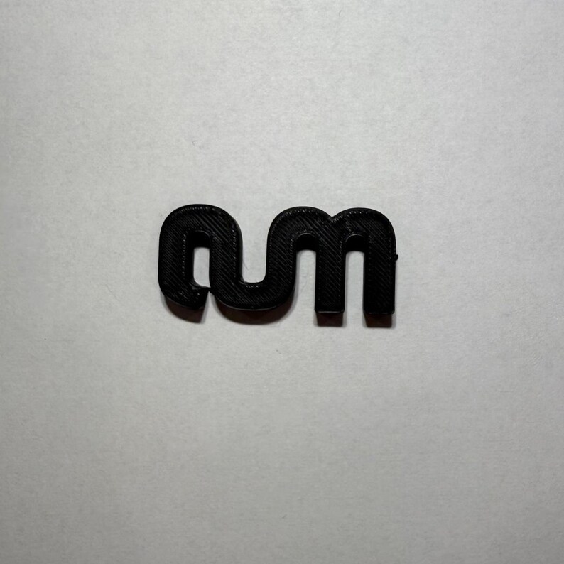 May include: A black, three-dimensional plastic object shaped like the lowercase letters 'nm' connected together. The surface of the object displays a visible, layered texture consistent with 3D printing, featuring a uniform matte black finish throughout.