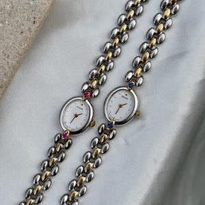 May include: Two Seiko Tisse wristwatches with oval white faces and gold-tone hands. The bands feature a two-tone silver and gold link design. One watch has pink accent stones, while the other features blue accent stones at the top and bottom lugs.
