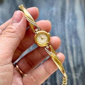 Può includere: Un orologio da polso dorato con quadrante color crema e lancette dorate. L'orologio ha un cinturino dorato e argentato con accenti decorativi e piccole pietre preziose. Un accessorio classico ed elegante.