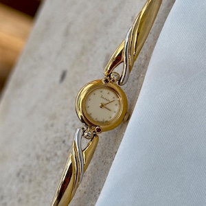 May include: A two-tone gold and silver-colored wristwatch with a round cream-colored dial featuring the brand name Jano. The watch band consists of interlocking gold and silver-colored metal links, accented with small clear stones and red gems.