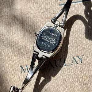 May include: Silver-toned wristwatch with a rectangular face and a delicate chain band. The watch face is marked with the text "SOMETHING" and "SOM-101". The watch is displayed on a textured, neutral-toned surface.