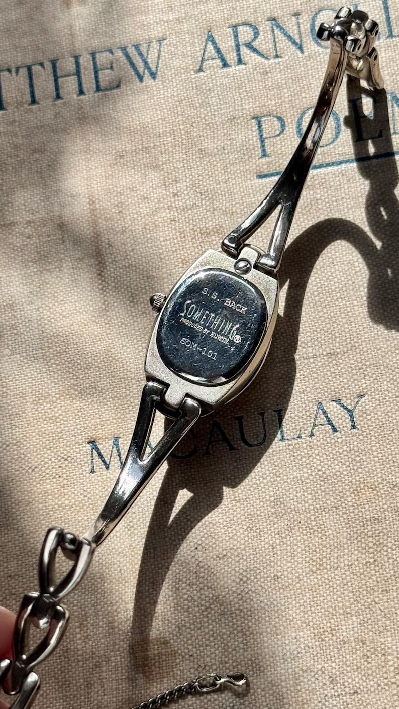 May include: Silver-toned wristwatch with a rectangular face and a delicate chain band. The watch face is marked with the text "SOMETHING" and "SOM-101". The watch is displayed on a textured, neutral-toned surface.