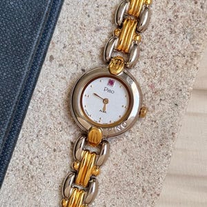 Puede incluir: Un reloj de dos tonos dorado y plateado con una esfera blanca y una joya rosa en la posición de las 12 en punto. La esfera del reloj dice "Pino" y "Alba" y tiene un pequeño segundero. El reloj tiene una correa de eslabones.