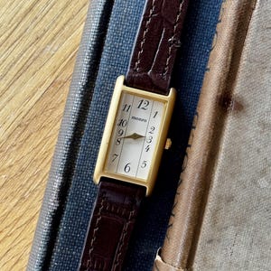 May include: A rectangular wristwatch with a gold-tone case and a white dial featuring black Arabic numerals from 1 to 12 and the text monro. The watch has a dark brown leather strap with a textured finish and gold-tone hands for the time display.