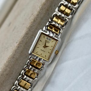 May include: A rectangular Seiko Quartz Phoenix wristwatch with a gold-toned textured dial, two small clear stones at the twelve o'clock position, and a two-tone silver and gold metal link bracelet with a polished finish and a side crown.