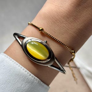 May include: Silver-tone bracelet with an oval, yellow-green gemstone center. A delicate gold chain bracelet is also visible. The jewelry is worn on a wrist, against a white shirt.