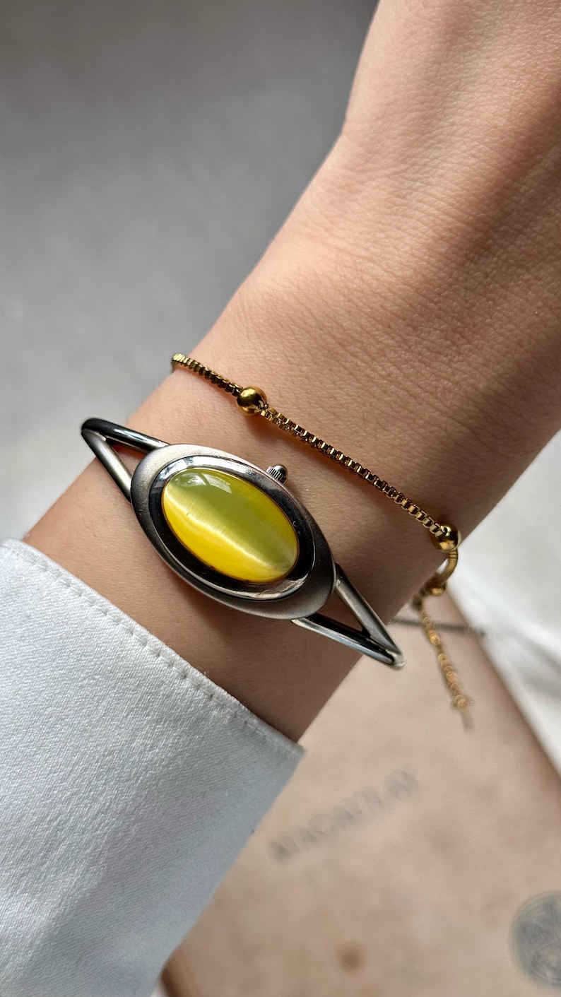 May include: Silver-tone bracelet with an oval, yellow-green gemstone center. A delicate gold chain bracelet is also visible. The jewelry is worn on a wrist, against a white shirt.