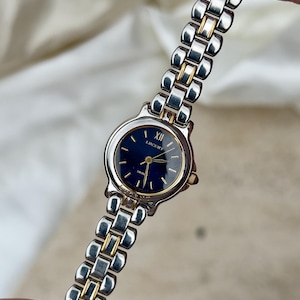May include: A two-tone wristwatch featuring a round case with a dark blue dial, gold-tone hands, and a Roman numeral at the twelve o'clock position. The word LUCENT is printed on the dial. The band consists of alternating silver and gold-tone links.