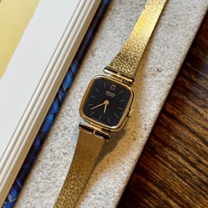 May include: A gold-tone Seiko Quartz wristwatch featuring a square case with rounded corners and a black dial. The watch has thin gold-tone hands and a textured gold-tone metal link bracelet with a clasp closure. The dial displays the Seiko logo.