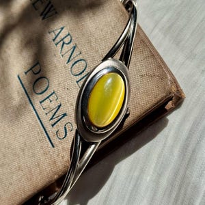 May include: A silver bracelet with an oval, yellow gemstone center. The bracelet is resting on a vintage book titled "Matthew Arnold Poems". The book has a worn, tan cover. The bracelet's design includes a chain link closure.