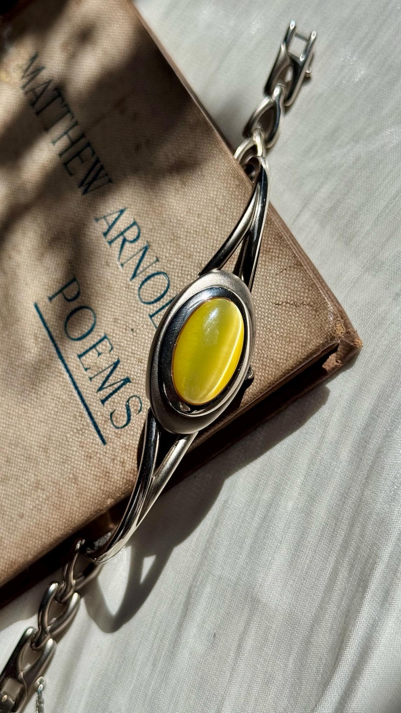 May include: A silver bracelet with an oval, yellow gemstone center. The bracelet is resting on a vintage book titled "Matthew Arnold Poems". The book has a worn, tan cover. The bracelet's design includes a chain link closure.