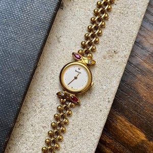 May include: A gold-tone wristwatch featuring a round white dial with black hands and markers. The watch band consists of a linked chain design. Two marquise-cut red stones and two small clear stones are set at the top and bottom of the watch case.