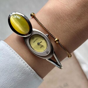 May include: A silver-toned wristwatch with a pale yellow face and a matching oval-shaped stone. The watch features the word "SOMETHING" on the face. A delicate gold-toned bracelet with small beads is also visible.