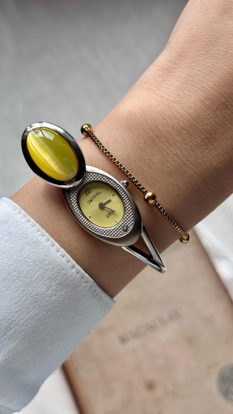 May include: A silver-toned wristwatch with a pale yellow face and a matching oval-shaped stone. The watch features the word "SOMETHING" on the face. A delicate gold-toned bracelet with small beads is also visible.