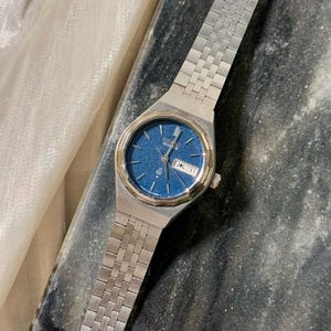 May include: A silver Seiko Quartz watch with a blue face. The watch has a rectangular dial with a date display and a silver link strap. The watch is shown on a dark, textured surface.