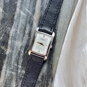 May include: A rectangular wristwatch with a white face and gold-tone detailing. The watch features a black leather strap. The watch face displays the word "AUREOLE". The watch is a quartz movement.