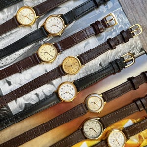 May include: A collection of ten analog wristwatches arranged in rows, featuring round cases in gold and silver tones with white or metallic dials. The watches are equipped with leather straps in black and brown, some with textured finishes and gold buckles.
