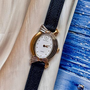 May include: A GRES quartz watch with a white oval face, gold and silver-toned casing, and a dark blue leather strap. The watch face displays the brand name and the word "QUARTZ".