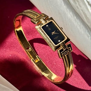 Puede incluir: Un reloj de pulsera dorado con una esfera negra rectangular y agujas doradas. El reloj forma parte de una pulsera de brazalete dorada. La esfera del reloj tiene la marca "Givenchy Paris" y dos pequeños diamantes. El reloj está sobre un fondo rojo.