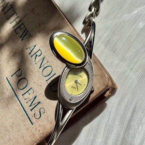 May include: A silver-toned wristwatch with a yellow face and a matching oval-shaped stone. The watch features a chain-link band and is set against a book titled "Matthew Arnold Poems."