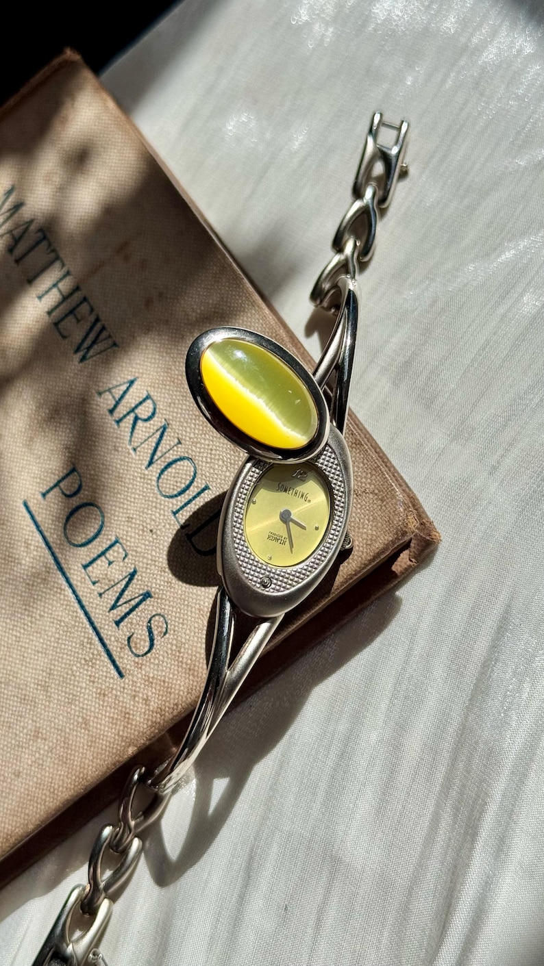 May include: A silver-toned wristwatch with a yellow face and a matching oval-shaped stone. The watch features a chain-link band and is set against a book titled "Matthew Arnold Poems."