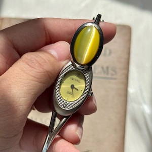 May include: A vintage silver-toned watch with a hinged cover and a yellow-green stone. The watch face is yellow with the word "SOMETHING" and the brand name "EDWIN" printed on it. The watch has a bangle-style band.