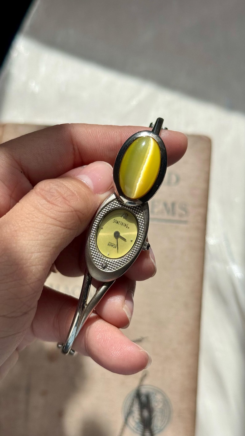May include: A vintage silver-toned watch with a hinged cover and a yellow-green stone. The watch face is yellow with the word "SOMETHING" and the brand name "EDWIN" printed on it. The watch has a bangle-style band.