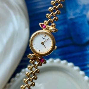 Könnte beinhalten: Goldfarbene Armbanduhr mit weißem Zifferblatt und filigranen Zeigern. Die Uhr hat ein goldenes Armband mit einem einzigartigen Kettendesign, verziert mit rosa und klaren Edelsteinen. Auf dem Zifferblatt steht das Wort "Inven" in Schreibschrift.