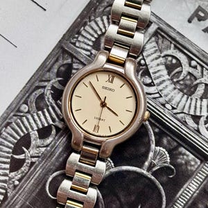 Puede incluir: Un reloj Seiko Lucent plateado y dorado con esfera color crema. El reloj presenta detalles dorados en la correa y alrededor de la esfera. Los números romanos marcan las posiciones de las 12 y las 10 en punto. La correa del reloj está hecha de eslabones plateados y dorados.