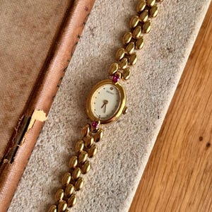 Puede incluir: Un reloj Seiko de tono dorado con esfera ovalada, fondo blanco y brazalete de eslabones. El reloj presenta un pequeño diamante en la posición de las doce y dos piedras rosas engastadas en la parte superior e inferior de la caja.