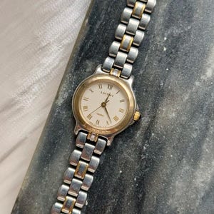 Puede incluir: Un reloj de cuarzo Lucent plateado y dorado con esfera blanca y números romanos. El reloj tiene un bisel dorado y una correa de eslabones plateada y dorada. El reloj está sobre una superficie gris oscuro.