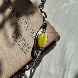 May include: A silver bracelet with a large oval yellow stone is displayed on a vintage book titled "Matthew Arnold Poems". The bracelet has a chain and a clasp. The book is tan with blue text.