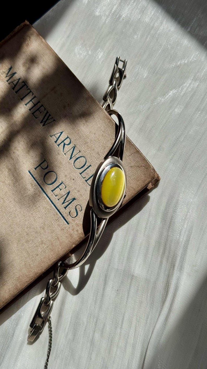 May include: A silver bracelet with a large oval yellow stone is displayed on a vintage book titled "Matthew Arnold Poems". The bracelet has a chain and a clasp. The book is tan with blue text.