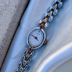 May include: A two-tone wristwatch with a silver and gold-colored link bracelet. The oval watch face is white with the text TISSÉ and JAPAN printed on it. Two small pink stones are set at the top and bottom where the bracelet meets the case.