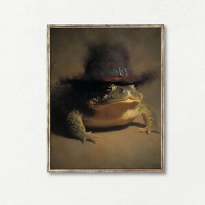 May include: A framed print depicting a frog wearing a dark hat with a buckle. The frog is rendered in detail with green and yellow skin. The artwork has a vintage, painterly style, set against a muted brown background.