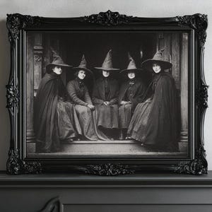May include: A monochrome framed print depicting five figures in witch hats and cloaks. The image is set within an ornate black frame. The figures are seated, creating a vintage, gothic aesthetic.