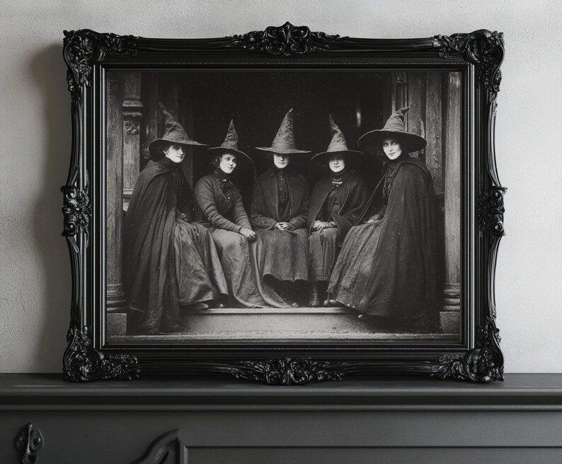 May include: A monochrome framed print depicting five figures in witch hats and cloaks. The image is set within an ornate black frame. The figures are seated, creating a vintage, gothic aesthetic.