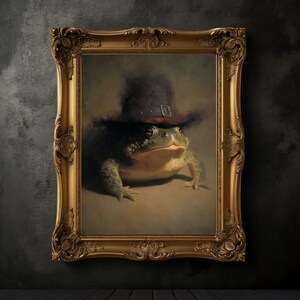 May include: A framed painting of a green frog wearing a dark hat with a buckle. The frog is centered in the ornate gold frame against a neutral background. The artwork has a classic, vintage aesthetic.