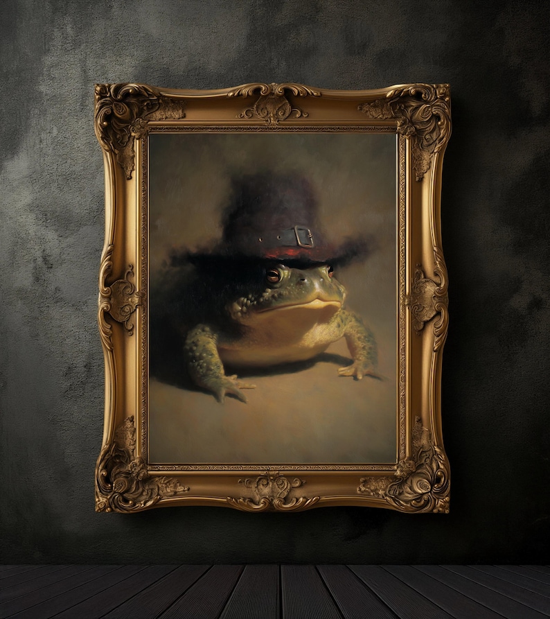 May include: A framed painting of a green frog wearing a dark hat with a buckle. The frog is centered in the ornate gold frame against a neutral background. The artwork has a classic, vintage aesthetic.