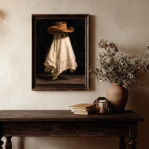 Pu&ograve; includere: Una stampa d'arte incorniciata raffigurante una figura spettrale drappeggiata in un lenzuolo bianco e con un cappello da cowboy marrone su sfondo scuro. L'opera &egrave; in una cornice di legno scuro, appesa a una parete sopra un tavolo in legno.