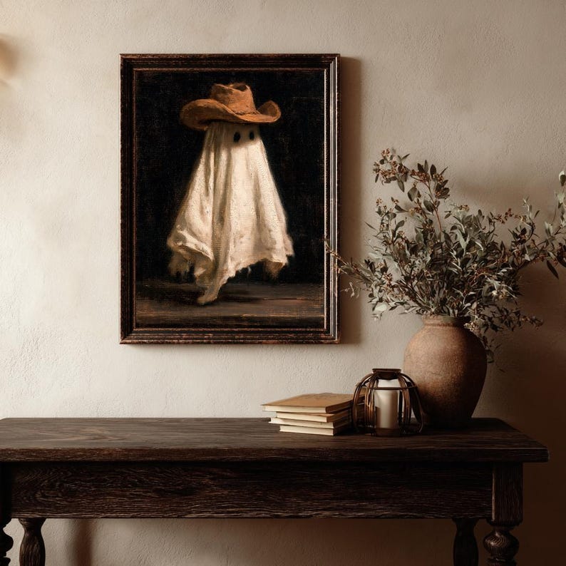 Pu&ograve; includere: Una stampa d'arte incorniciata raffigurante una figura spettrale drappeggiata in un lenzuolo bianco e con un cappello da cowboy marrone su sfondo scuro. L'opera &egrave; in una cornice di legno scuro, appesa a una parete sopra un tavolo in legno.