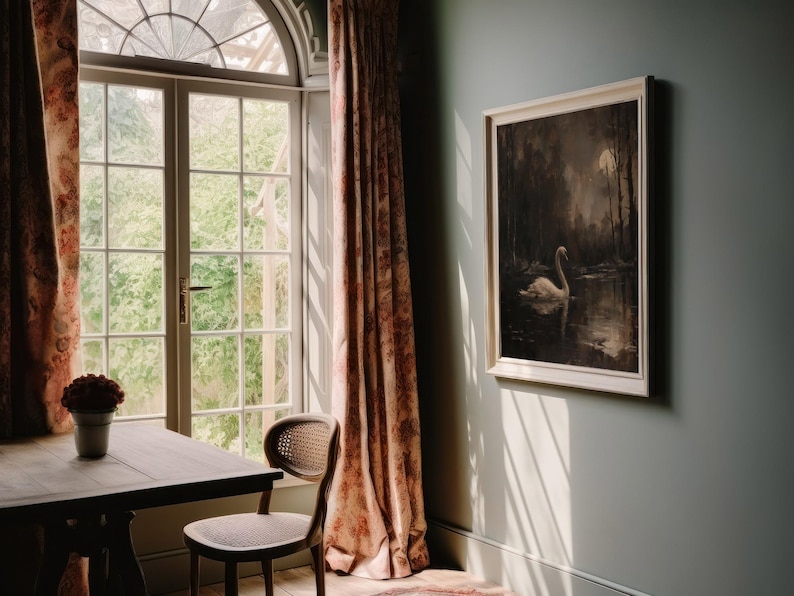 May include: A vintage-style framed print of a white swan swimming in a dark, moonlit lake. The print is hanging on a light green wall in a room with a window and a table with a chair.