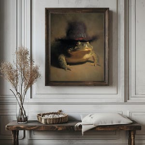 May include: A framed artwork shows a green frog wearing a black hat with a red band. The frog is centrally positioned, against a neutral backdrop. The frame is dark brown, and the artwork is displayed on a white wall.