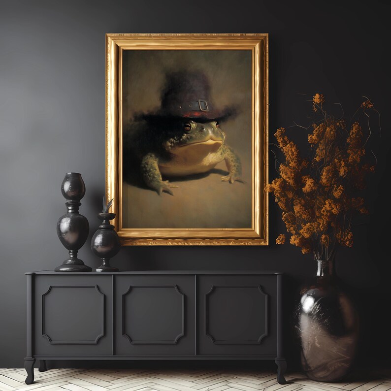 May include: A framed painting of a frog wearing a hat. The artwork has a gold frame and is displayed above a black cabinet. Decorative vases and dried flowers are also visible.