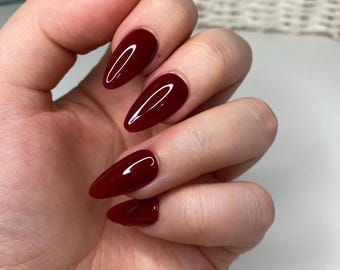 Op de afbeelding: Close-up van een hand met lange, amandelvormige nagels die in een diep rood zijn gelakt.