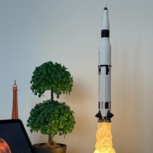 May include: A Saturn V rocket lamp with a glowing flame effect. The lamp is white with black accents and the text "SATURN V" and "NASA" on the base. A small green plant and a tablet are in the background.