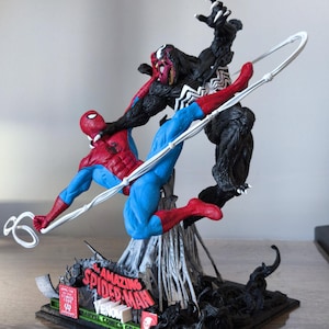 May include: A detailed figurine depicting Spider-Man in a dynamic pose, battling Venom. Spider-Man is in a red and blue suit, while Venom is a black figure with white accents. The base features the words "Amazing Spider-Man" and "Marvel Comics Group."
